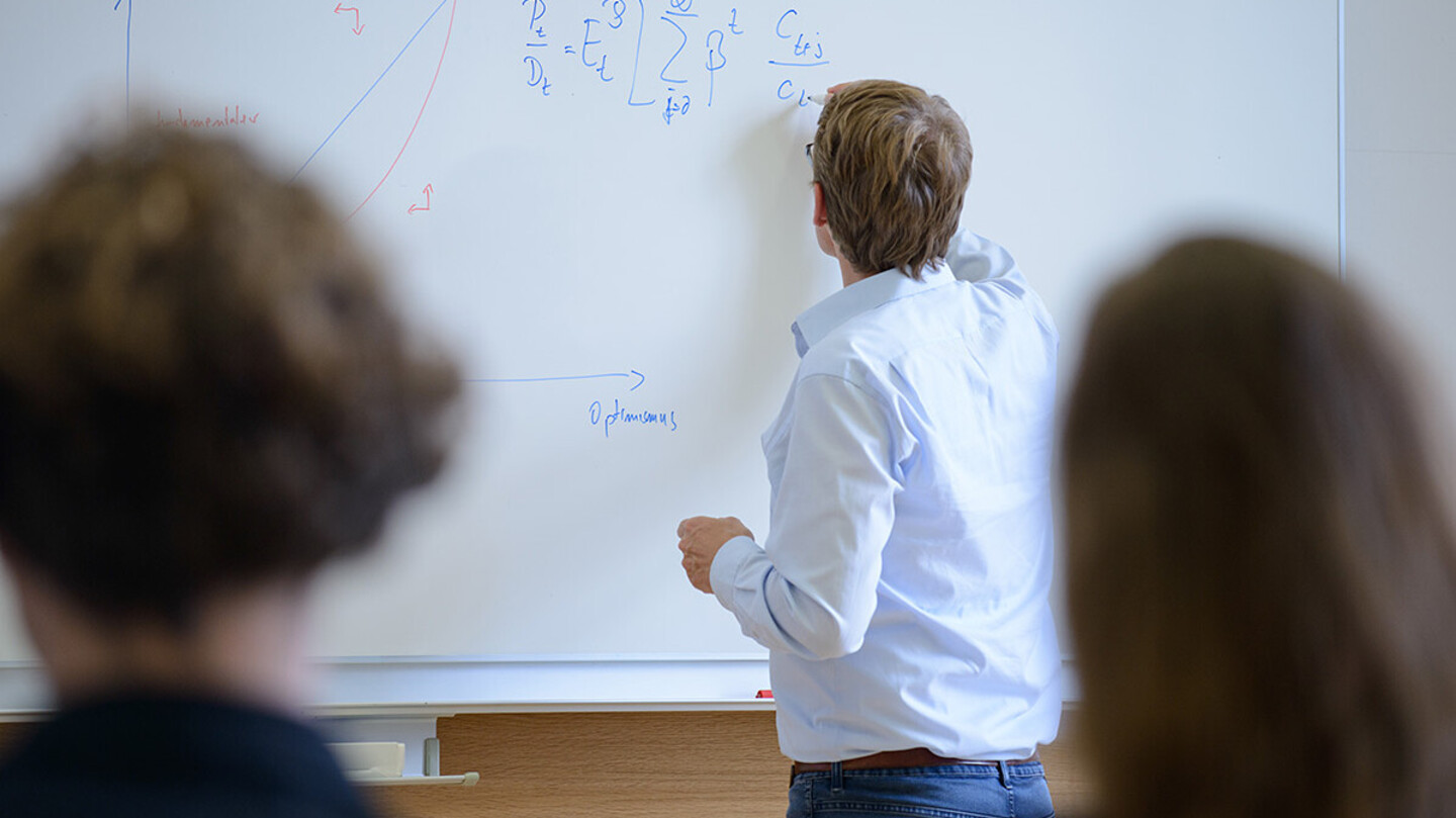 Zwei Personen schauen zu, wie ein Mann eine Formel auf ein Whiteboard aufschreibt.