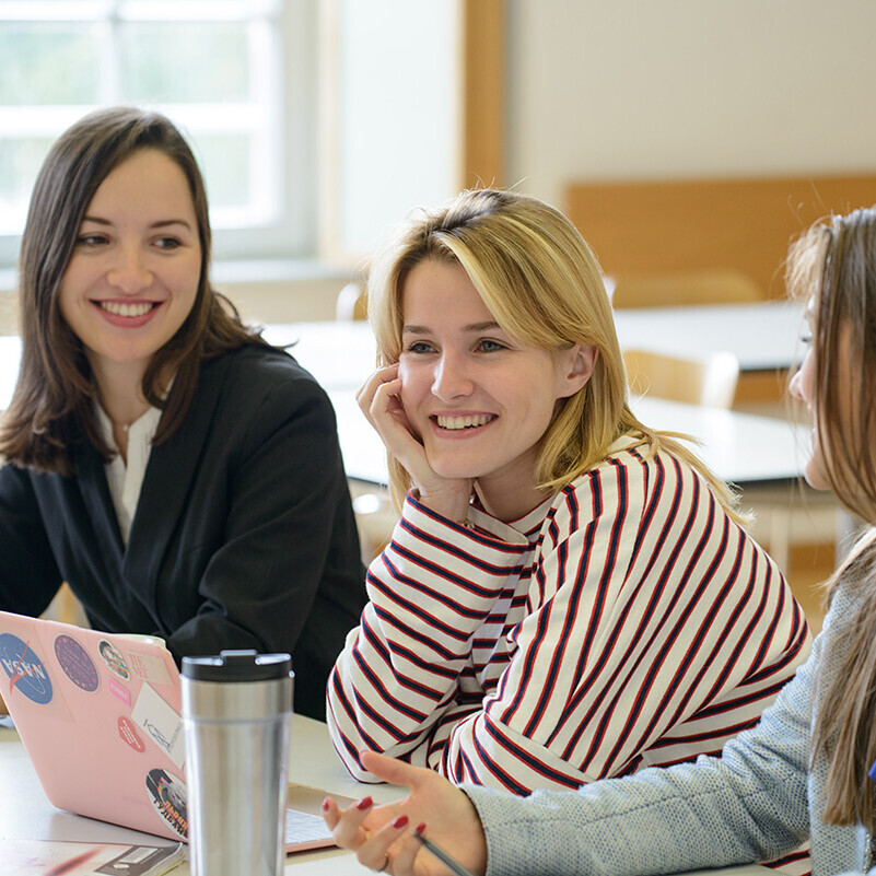 Drei Studierende sitzen lächelnd in Seminarraum