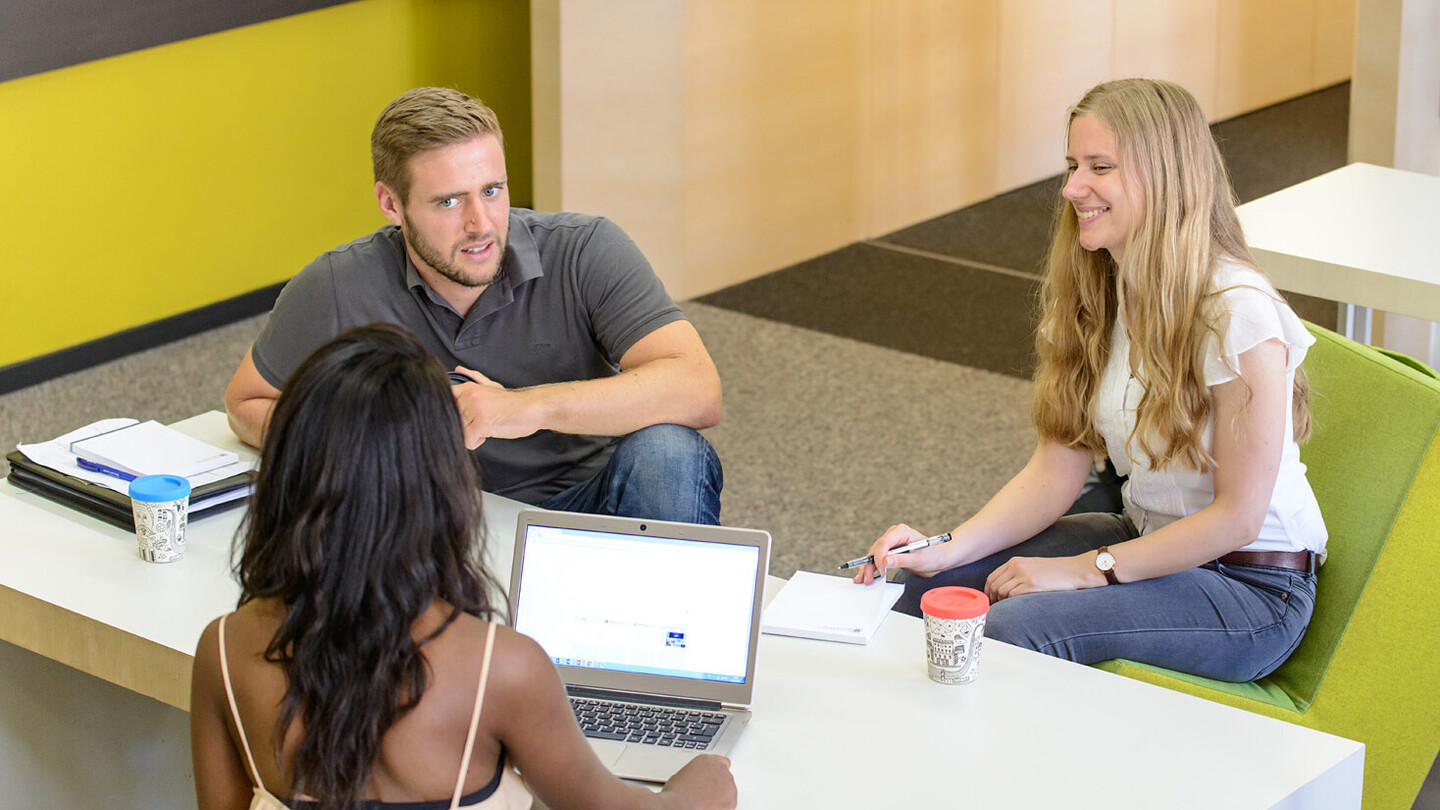 Zwei Studentinnen und ein Student sitzen gemeinsam mit Lernmaterialien an einem Tisch und unterhalten sich.