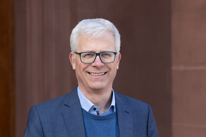 Portrait von Eckhard Janeba. Er trägt ein hellblaues Hemd, einen blauen Pullover sowie einen blauen Blazer und steht lächelnd vor einem Gebäude.